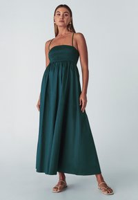 Abito lungo verde scuro, con corpetto arricciato e spalline sottili, caratterizzato da una gonna fluttuante e una texture morbida. Sandali piatti visibili.