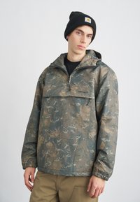 Svetrová bunda s camouflage vzorom a polovičným zipsom, kapucňou a elastickými manžetami, vyrobená z vodoodolného materiálu. Čierny logo detail.