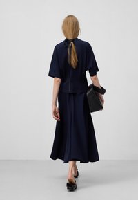 Robe bleu marine, longueur genou, avec des manches courtes et un détail noué dans le dos, associée à un sac à main noir et des chaussures plates, vue de derrière.