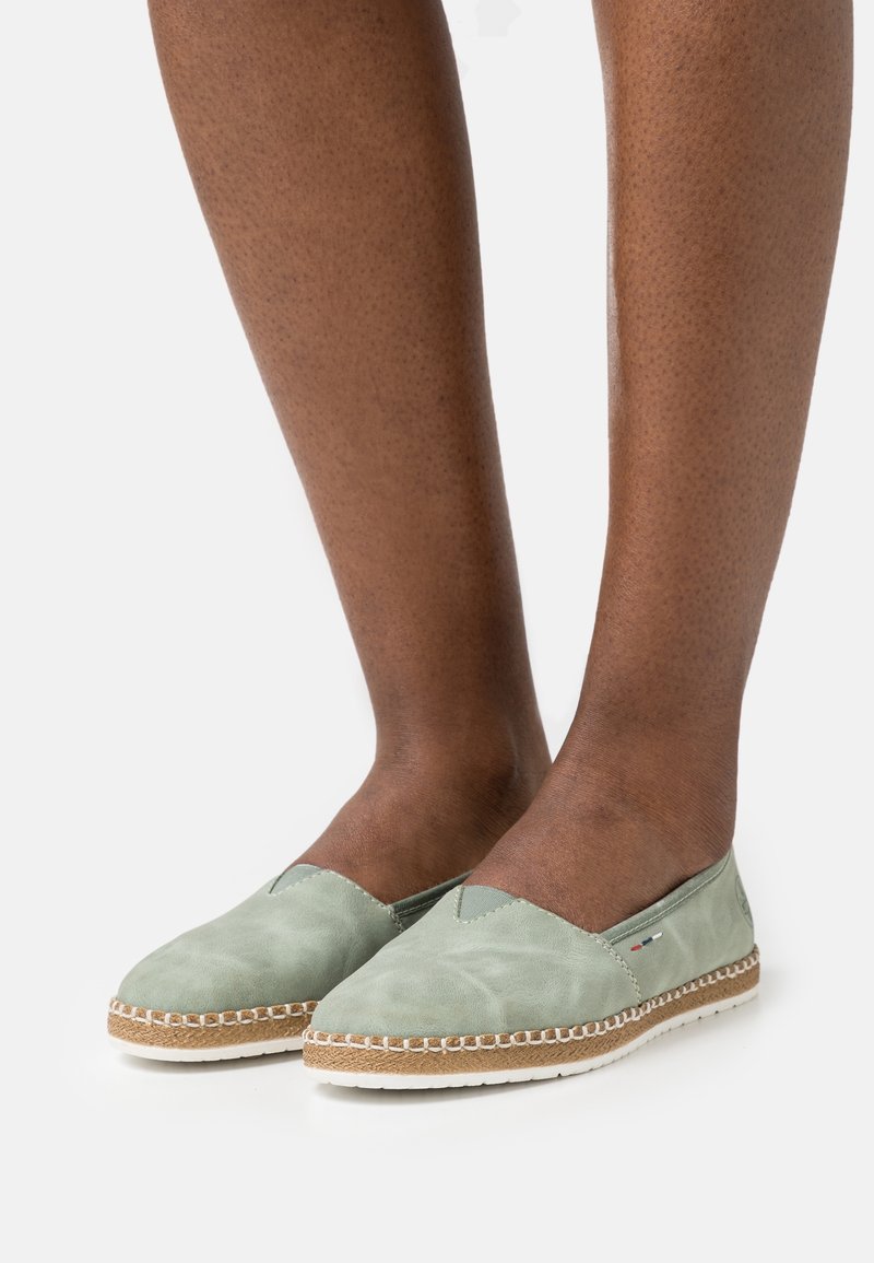 Pieds portant des espadrilles légères vert clair à enfiler avec des coutures blanches et des semelles à détail corde sur fond blanc.