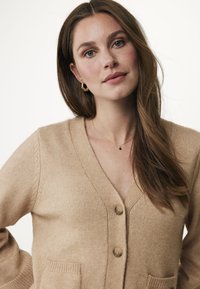 Beige cardigan med V-ringning, med två framfickor och knappar av trä. Mjuk textur, ribbade ärmslut och avslappnad passform.