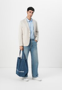 Beige Blazer, hellblaues Hemd und lockere Jeans. Dunkelblaue Umhängetasche mit weißem Text. Weiße Sneakers vor einem schlichten Hintergrund.
