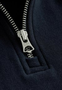 Kovový zips na tmavomodrom texte s nápisom "SUPER DRY" vyrytým na ťahadle, čiastočne rozopnutý, odhaľujúci čierne pozadie.