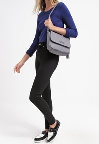 Vrouw in zwarte skinny jeans, blauwe langermige top, zwart-witte loafers, die een grijze schoudertas draagt op een witte achtergrond.
