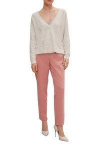 Pull en maille beige clair avec un col en V profond, associé à un pantalon rose sur mesure et des chaussures blanches à talons hauts, créant un look superposé.