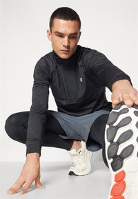 Haut à manches longues zippé noir, shorts gris et leggings noirs. Chaussures de sport blanches avec semelle texturée. Modèle en position d'étirement.