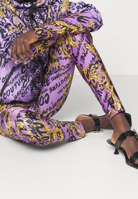 Lila leggings med svarta och guld grafiska tryck, med utsmyckade mönster. Bärs med svarta remsandaler, i sittande pose med korslagda ben.