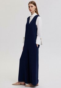 Jūras zila kombinezons ar V veida kaklu, platajām kājām un sānu kabatām, uzlicis virs balta blūzes ar pogām pie aprocēm un gludu apdari.