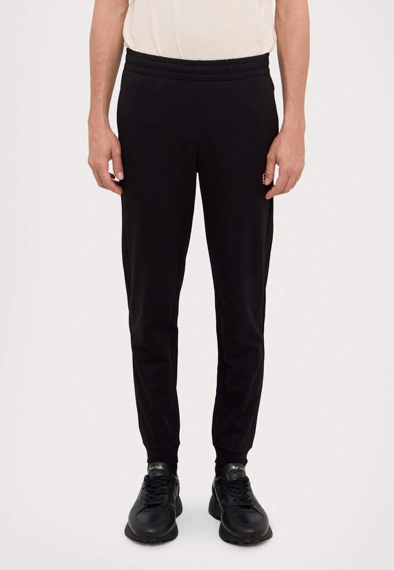 Fekete melegítőnadrág, rugalmas derékkal, slim fit szabással és bordázott szegéllyel. Fekete sportcipővel párosítva, amelynek textúrázott talpa van.