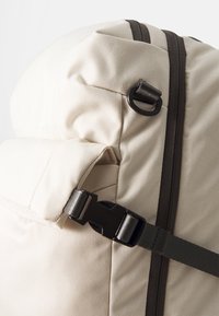 Beige ryggsekk med avrundet topp og mørkegrå detaljer. Har en plastlås og glidelås, laget av slitesterkt, strukturert stoff.