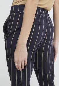 Marine gestreifte Hose mit goldenen vertikalen Linien, ausgestattet mit einem hohen Bund, Seitentaschen und einer eleganten Stofftextur. Die Hand ruht auf der Hüfte.