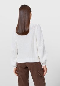 Bílý pletený svetr s texturovaným vzorem, žebrovanými manžetami a lemem. Zadní část má volnější střih a široký výstřih. Spárováno s hnědými kalhotami.