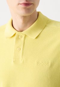 Gros plan sur une personne portant un polo jaune avec un col côtelé, deux boutons et "BOSS" brodé sur la poitrine.