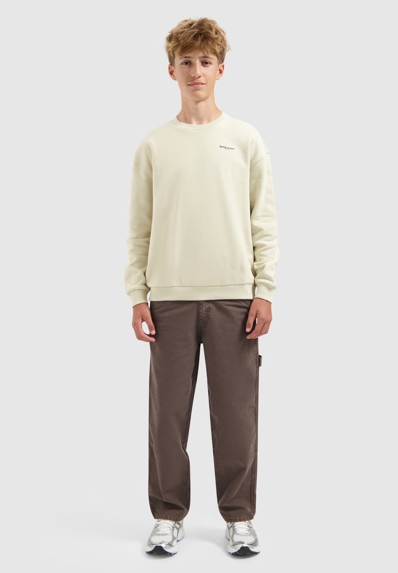 Tienerjongen staat, gekleed in een beige sweatshirt, bruine broek en witte sneakers tegen een effen lichtgrijze achtergrond.