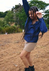 Femme aux longues tresses portant une chemise bleue et une jupe beige danse joyeusement dans un champ de terre avec des arbres verts en arrière-plan.