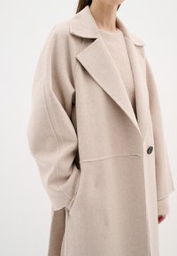 Cappotto in misto lana beige con colletto a revers, vestibilità oversize e un singolo bottone nero. Texture morbida con tasche laterali e spalle scese.