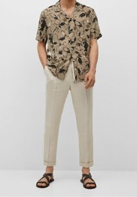Chemise à manches courtes beige avec des motifs abstraits bruns et noirs ; associée à un pantalon à jambes fuselées beige clair et des sandales foncées.