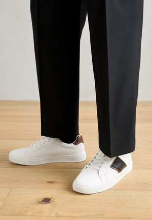 Weiße Sneakers mit glatter Textur, versehen mit braunen Lederakzenten und schwarzen Details, kombiniert mit schwarzen maßgeschneiderten Hosen. Hintergrund aus Holzboden.