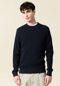 Marinblå stickad tröja med en texturerad yta, rund halsringning och ribbade muddar och fåll. Bärs över en vit T-shirt, minimalistisk design.