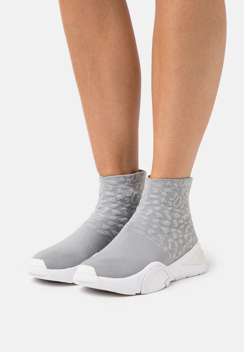 Enkelhoge grijze gebreide sokken-sneakers met subtiel gestructureerd patroon en dikke witte zool, gedragen op blote benen tegen een effen achtergrond.