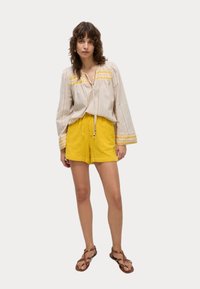 Blusa ligera beige con bordado amarillo y un cordón ajustable, combinada con pantalones cortos amarillos. Sandalias de tiras marrones completan el conjunto.