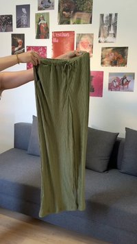 Personne tenant un pantalon large côtelé vert olive dans un salon avec un canapé gris et une œuvre d’art colorée sur le mur derrière.