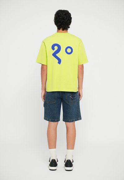 Neon green short-sleeve t-shirt featuring a large blue graphic of "20º" on the back, paired with dark denim shorts and black sneakers.