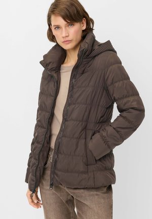Woman wearing a brown padded jacket with a high collar, beige top, and brown pants, standing with one hand in the jacket pocket.