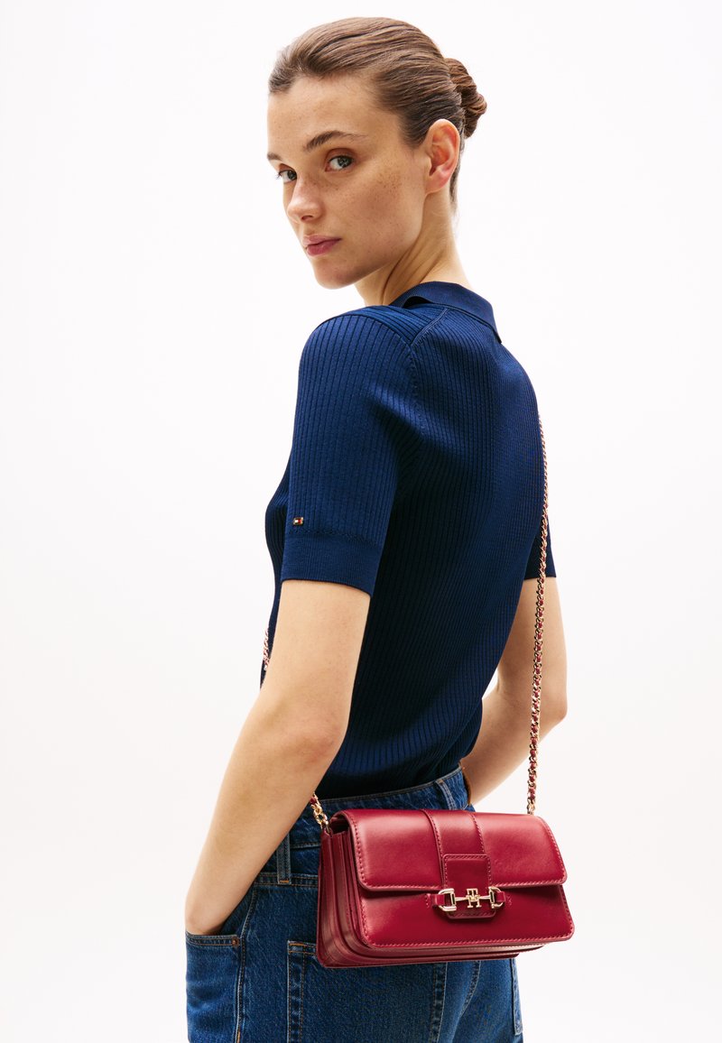 Navyblaues Rippstrickoberteil, kombiniert mit einer kleinen bordeauxroten Lederhandtasche, die einen goldenen Verschluss und eine Kettenriemen hat, vor einem weißen Hintergrund.