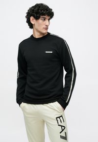 Vīrietis ar cirtainiem matiem, valkājot melnu garām piedurknēm džemperi ar baltām svītrām uz piedurknēm un bēšas krāsas bikses ar lielu EA7 logo.