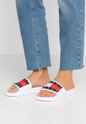 White slide sandals with a wide strap featuring a red and navy design and the text "TOMMY JEANS." Textured rubber sole, worn with blue jeans.