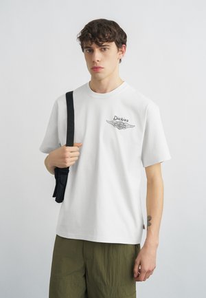 Jaunas vyras dėvi baltą Dickies marškinėlį, alyvuogių žalių kelnių ir juodą krepšio diržą per petį, stovintis priešais paprastą foną.