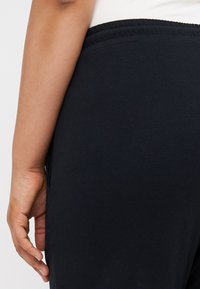 Černé elastické kalhoty z jemné tkaniny s pružným pasem. Detailní záběr zachycuje boční šev a ruku položenou poblíž boků. Měkká, hladká textura.