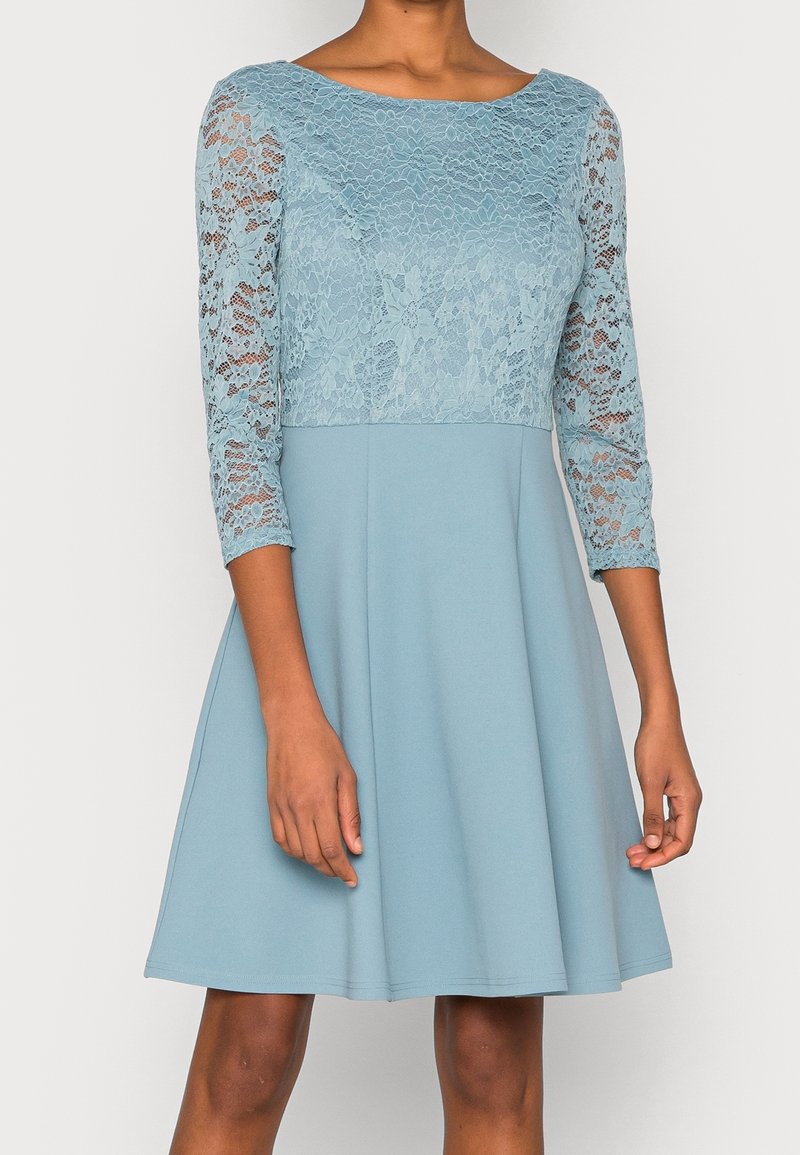 Une robe bleue avec un corsage en dentelle, des manches trois-quarts et une jupe évasée légère. Le tissu est lisse et doux.