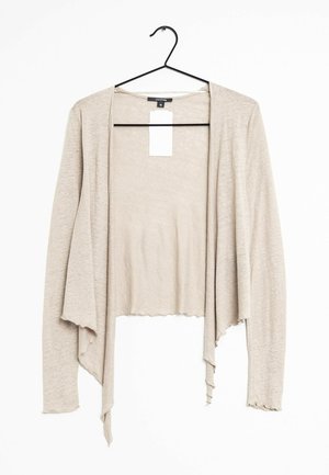Cardigan beige léger à manches longues avec ouverture devant, suspendu à un cintre noir avec une étiquette blanche de prix attachée au col.