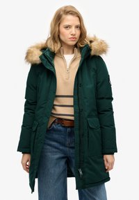 Abrigo acolchado verde oscuro con capucha forrada de pelo sintético, dos bolsillos frontales y cremallera delantera. Lleva puesto un suéter beige y unos vaqueros azules.