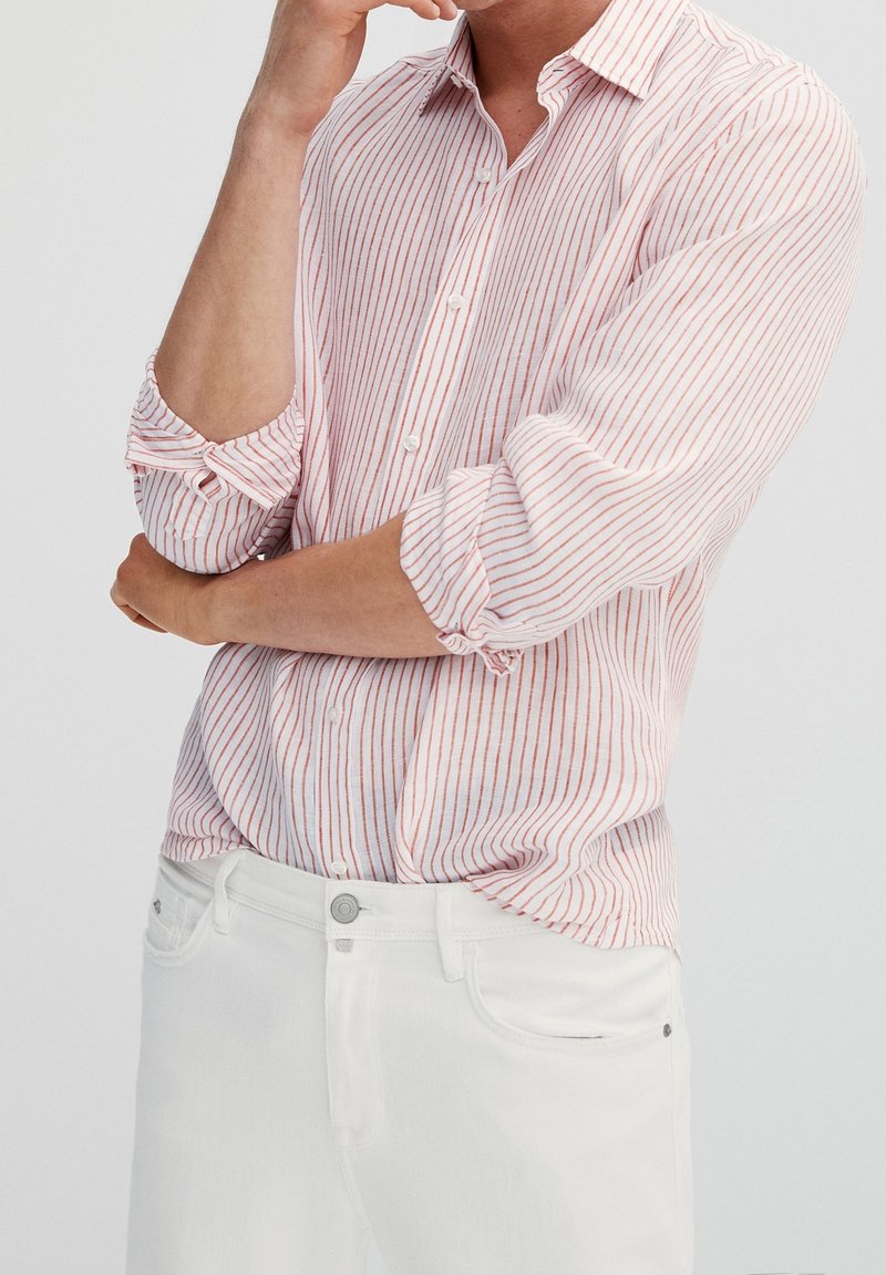 Homme portant un pantalon blanc et une chemise blanche à rayures verticales rouges, manches retroussées, main touchant le menton dans une pose réfléchie.