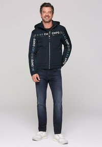 Navy zip-up jacket with hood, featuring printed accents in light blue. Paired with dark jeans and white sneakers. Smooth texture, tailored fit.