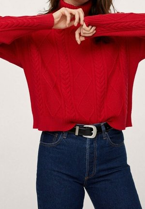 Donna che indossa un maglione rosso a trecce con maniche lunghe e collo alto, abbinato a jeans blu a vita alta e una cintura nera.