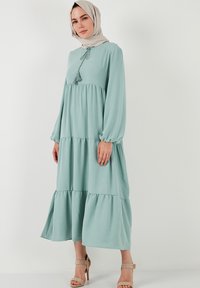 Light green, tiered maxi dress with long sleeves, tasseled neck detail, and soft texture. Accented with a subtle ruffle at the hem.