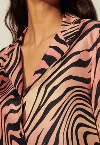 Chemisier en soie avec un schéma de couleurs rose et beige, arborant d'épaisses rayures zèbres noires, un col en V et une fermeture à boutons. Texture lisse.