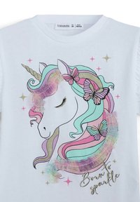 T-shirt bianco con una grafica di un unicorno con criniera arcobaleno pastello, accenti luccicanti, farfalle e la frase "Nato per brillare" in oro.