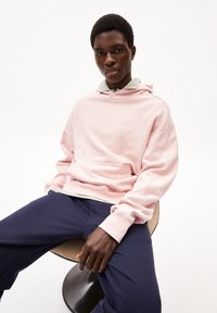 Felpa rosa con tasca frontale e logo ricamato, sovrapposta a una camicia grigia con colletto. Indossata con pantaloni blu, seduto su una sedia circolare.