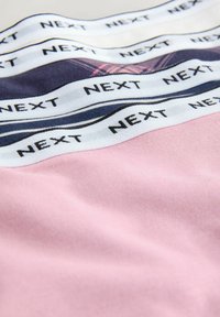 Pilas de ropa interior doblada con cinturillas blancas etiquetadas con "NEXT", con colores de tela en azul marino, cuadros y rosa.