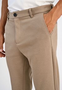 Calças bege com textura suave e tecido tecido, com bolsos laterais, fecho de botão único e ajuste sob medida.