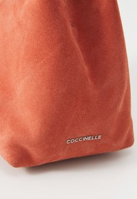 Raudona zomšinė rankinė su minkšta tekstūra, su sidabriniu iškilniu logotipu "COCCINELLE" šone ir paprastu, minimalistiniu dizainu.