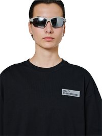 Osoba nosząca sportowe, lustrzane okulary przeciwsłoneczne oraz czarną, oversize'ową koszulę z małą naszywką "HALO 196819721981" na piersi.