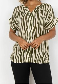 Blusa a maniche corte in tessuto beige con strisce a zebre verdi, caratterizzata da un collo a V e un fit rilassato. Abbinata a pantaloni neri.