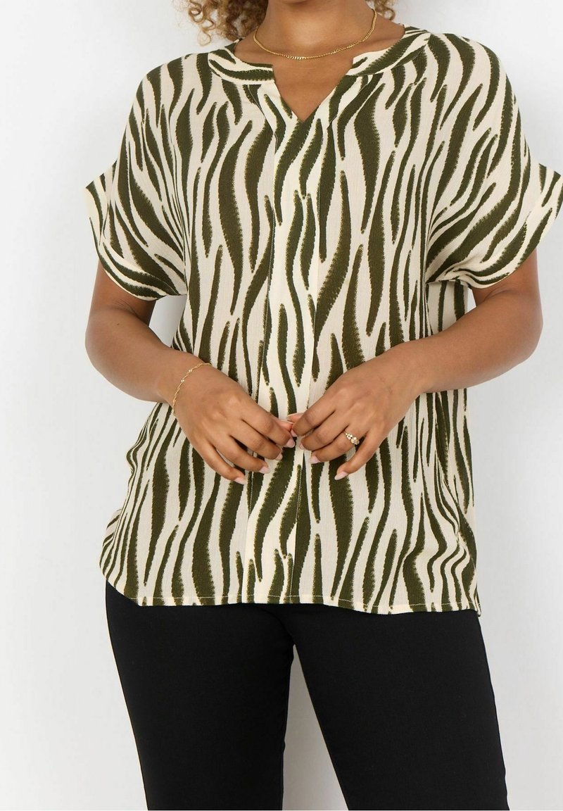Blusa a maniche corte in tessuto beige con strisce a zebre verdi, caratterizzata da un collo a V e un fit rilassato. Abbinata a pantaloni neri.