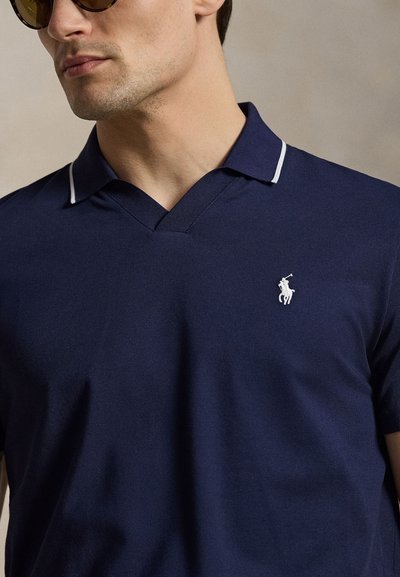 Άνδρας που φοράει σκούρο μπλε πόλο με λευκή επένδυση στο γιακά και λευκό κεντημένο λογότυπο παίκτη πόλο στο στήθος, με ορατά γυαλιά ηλίου.